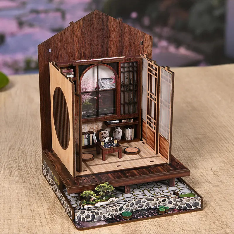 Fingers removing tatami mat piece from wooden sheet while assembling【Moss-Lantern Harmony Garden】， scattered miniature tea set, books, and bonsai tree parts on desk, immersive Japanese-style craft time