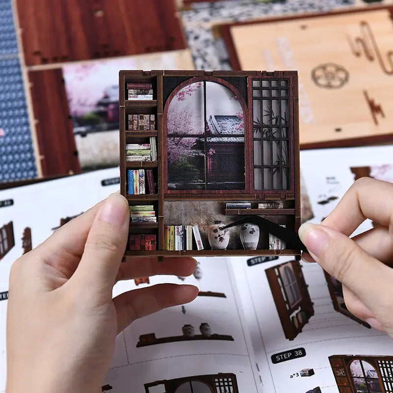 Two pairs of hands assembling【Moss-Lantern Harmony Garden】 on a bright desk, one using tweezers to insert miniature books into shelf, the other gluing shoji paper onto door frame grilles against the manual