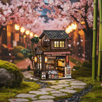 Kasuga Grocery Store DIY book nook miniature – Japanese two-story wooden grocery shop with warm amber LED lanterns, cherry blossom trees and stone path background, cozy Japanese street scene book nook kit
